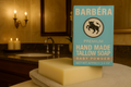 A close-up photograph features a light blue box of "BARBERA Premium Hand Made Tallow Soap" scented as "Baby Powder" in a well-appointed, softly lit bathroom setting. A large, unscented block of cream-colored soap rests on a light gray towel in the foreground, with the product packaging standing behind it. In the background, there is a dark marble countertop with a built-in sink, a large, dark-framed mirror, and a brass wall sconce with glowing candles, reflecting the warm atmosphere.